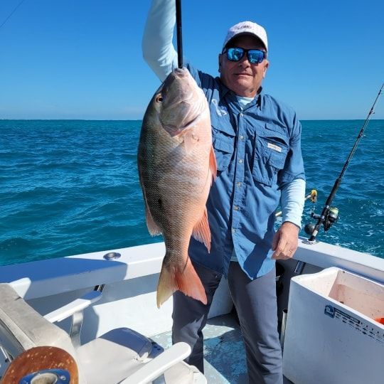 A person on a boat holds up a large red snapper fish against a bright blue ocean backdrop.