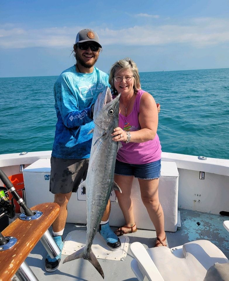 A man and a woman hold a large fish while standing on a boat on the ocean under a clear blue sky.