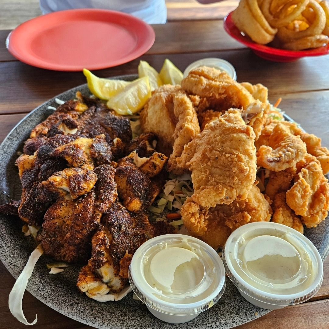 A platter of blackened and fried seafood with two dipping sauces, lemon wedges, and a side of curly fries.