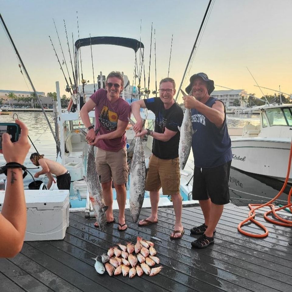 Three people holding large fish on a dock next to a boat, with a pile of smaller fish on the deck in front of them.