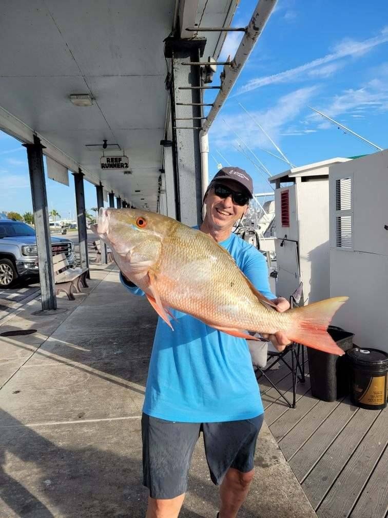 A person wearing a blue shirt and sunglasses holding a large, golden-hued fish on a dock.