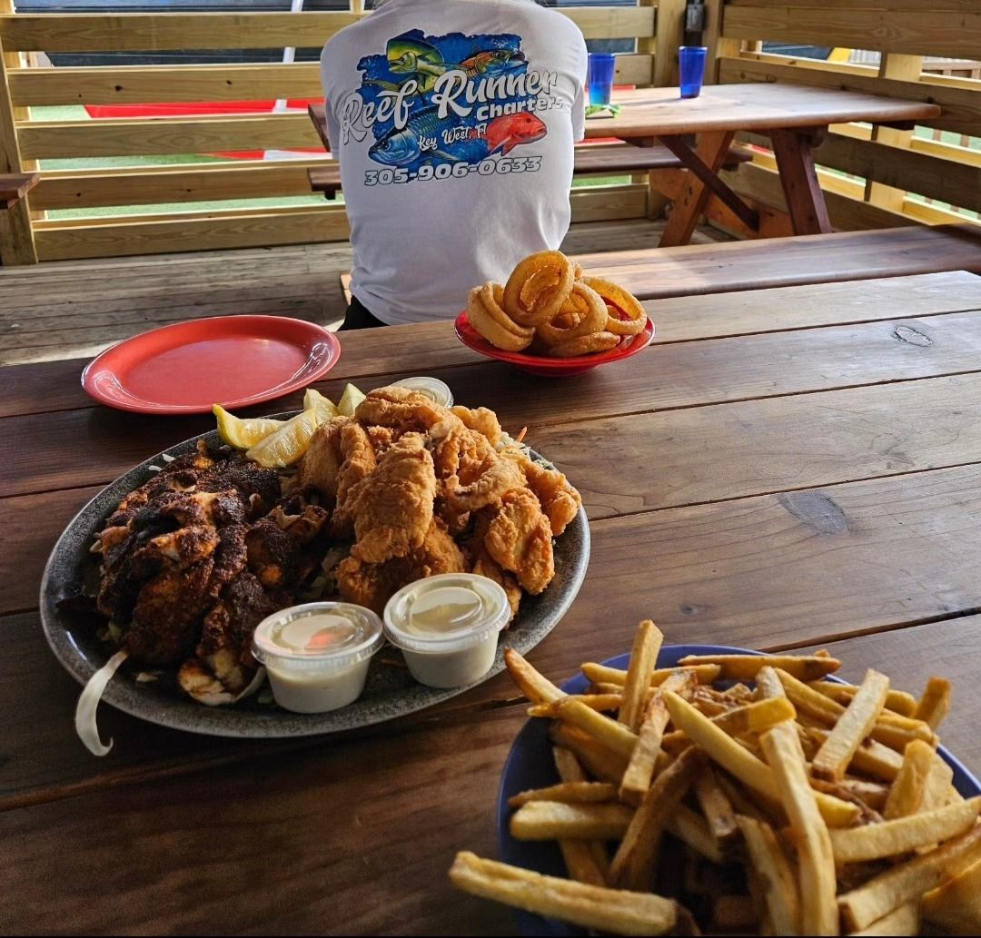 A large platter of fried seafood and fries sits on a wooden table, with a person wearing a graphic shirt in the background.
