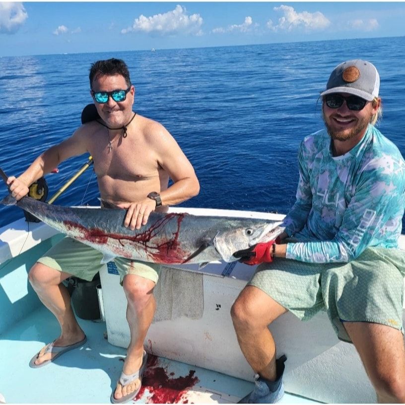 Two people on a boat holding a large, caught fish with visible blood against a blue ocean backdrop.