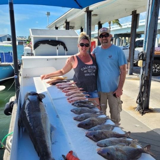 A couple stands on a dock behind a table displaying a large catch of fish, including one very large fish on the left.
