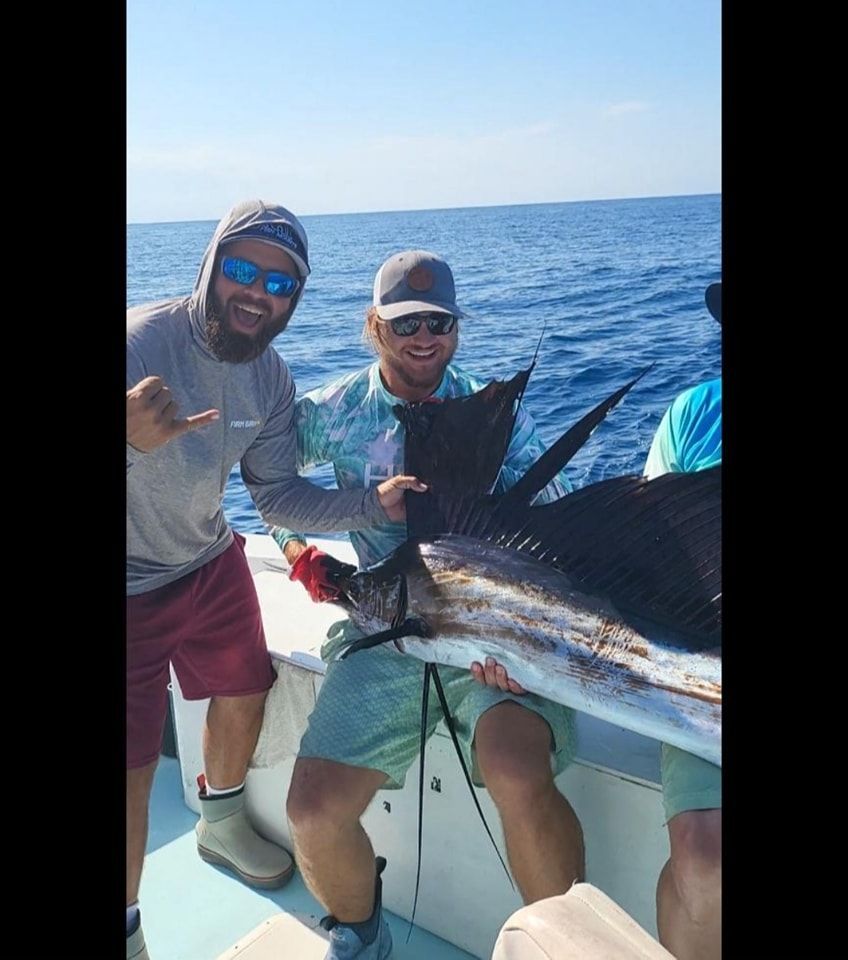 Two people on a boat holding a large sailfish with an extended dorsal fin, smiling under a bright blue sky.