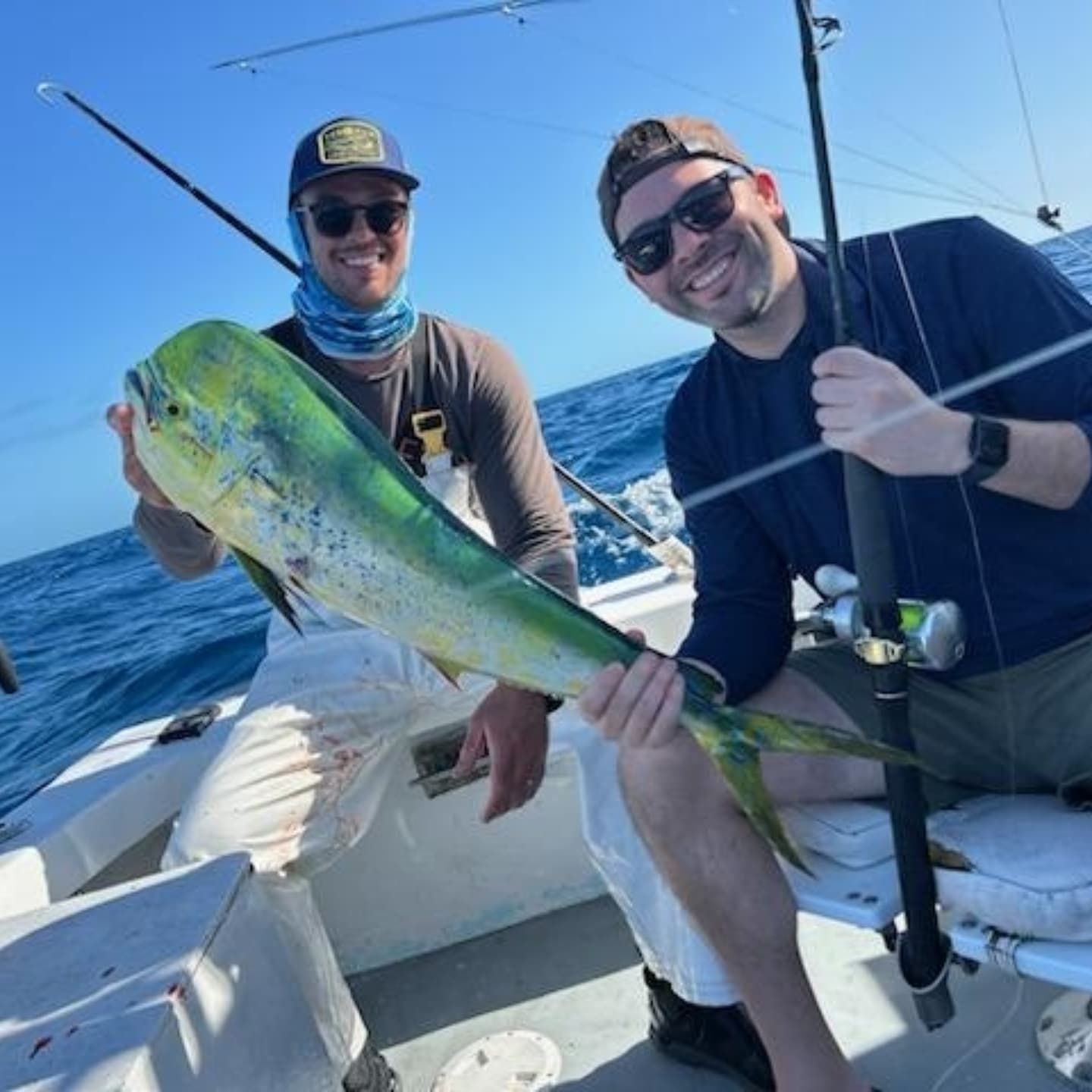 Two people in sunglasses and caps hold a colorful mahi-mahi fish on a boat on the ocean.