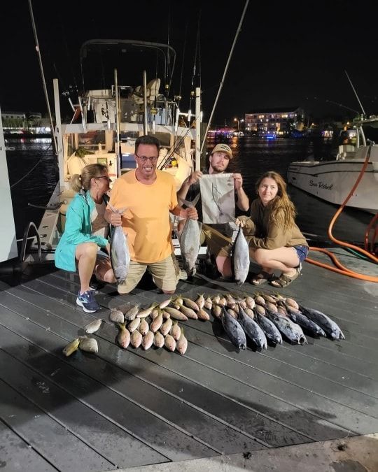 Four people pose on a dock at night next to a large haul of freshly caught fish and a fishing boat.