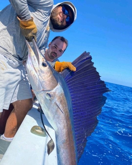 Two people on a boat hold up a large, silver sailfish with a prominent blue dorsal fin against a bright blue sky.