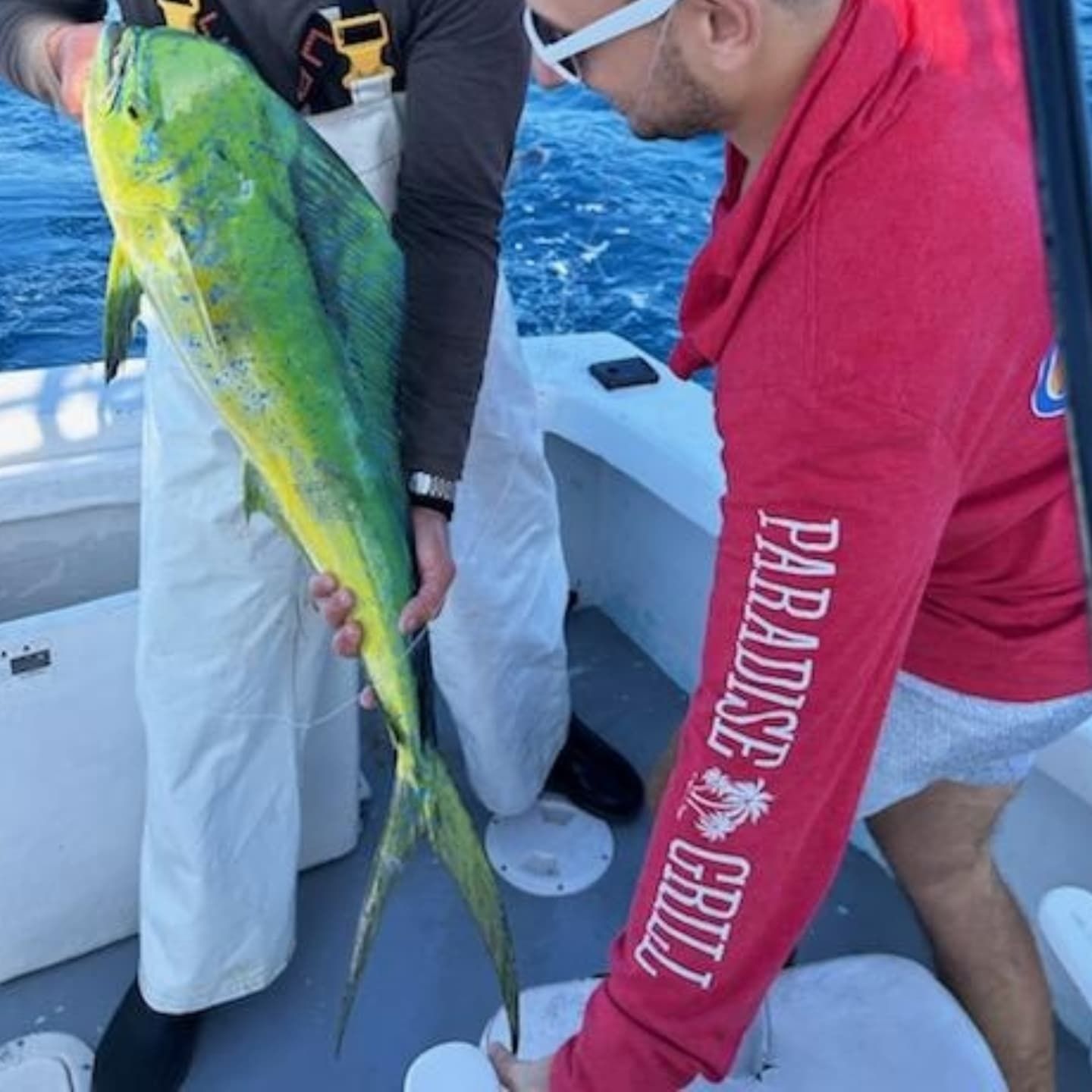 Two people on a boat hold a large, vibrant green and yellow mahi-mahi fish over the deck.
