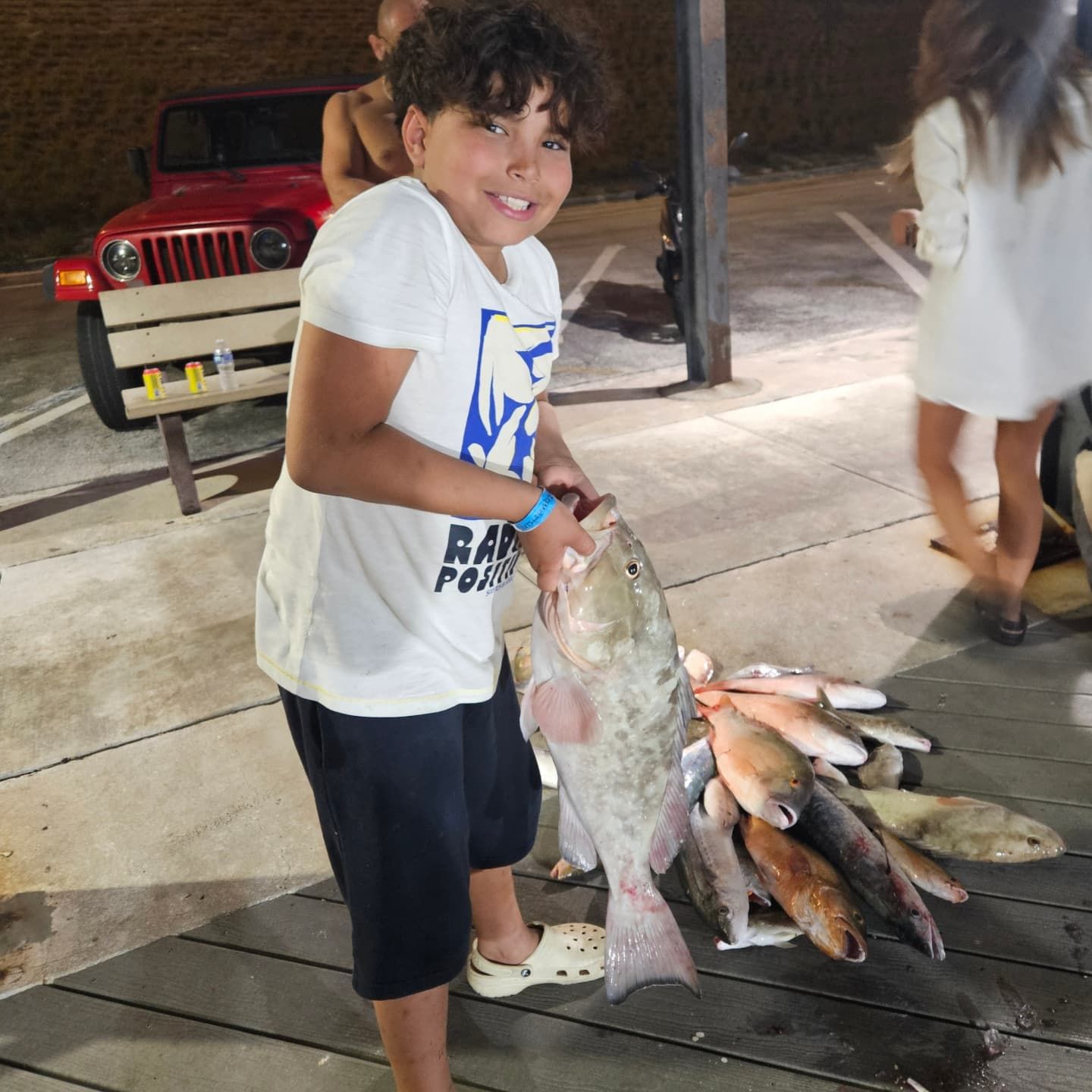A young person holding a large fish, standing next to a pile of other fish on a dock at night near a red jeep.
