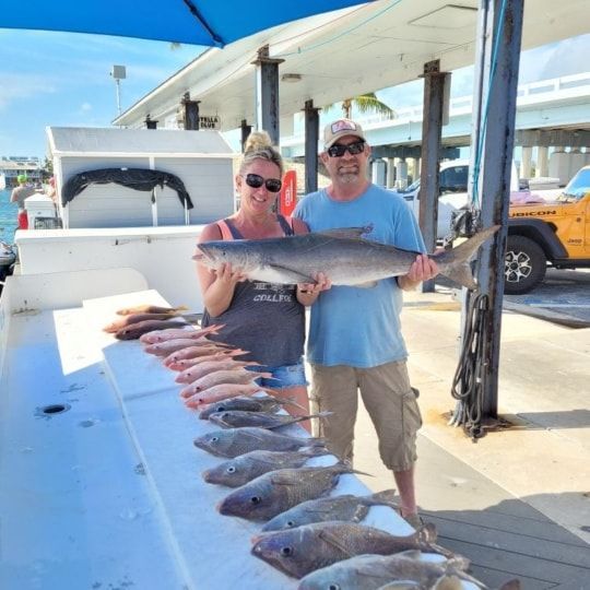 A couple holds a large fish while standing on a dock next to a table lined with several freshly caught fish.