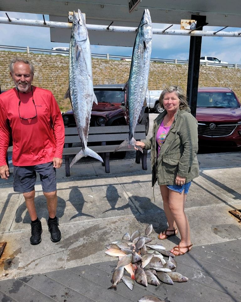 A person in a red shirt and a person in a green jacket stand outdoors with two large fish hanging and several on the ground.