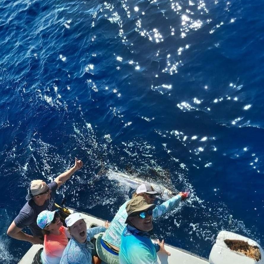 A group of people on a boat excitedly reach toward a billfish swimming in the deep blue, sunlit water.