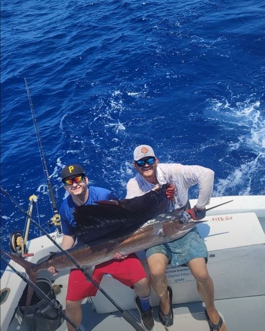 Two people holding a sailfish on a boat in blue ocean water, both wearing hats, sunglasses, and casual clothing.