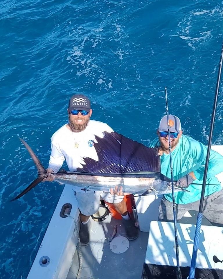 Two people on a boat holding a large sailfish with a prominent blue dorsal fin against a backdrop of deep blue ocean.