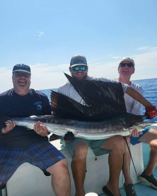 Three people smiling while sitting on a boat, holding a large sailfish with an extended dorsal fin against a blue sky.