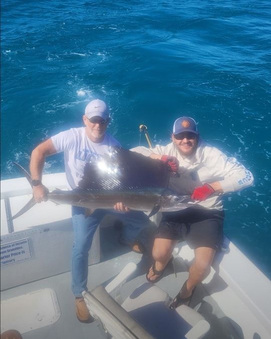 Two people on a boat holding a large sailfish up by the deck against a background of deep blue ocean.