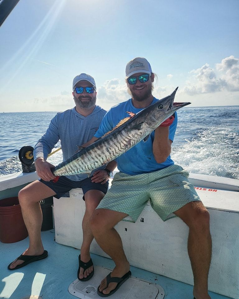 Two people sit on a boat deck, smiling and holding a long, striped wahoo fish caught at sea on a sunny day.