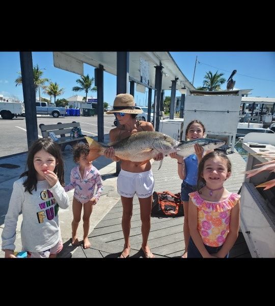 A person holding a large fish stands on a dock surrounded by children, with boats and palm trees in the background.