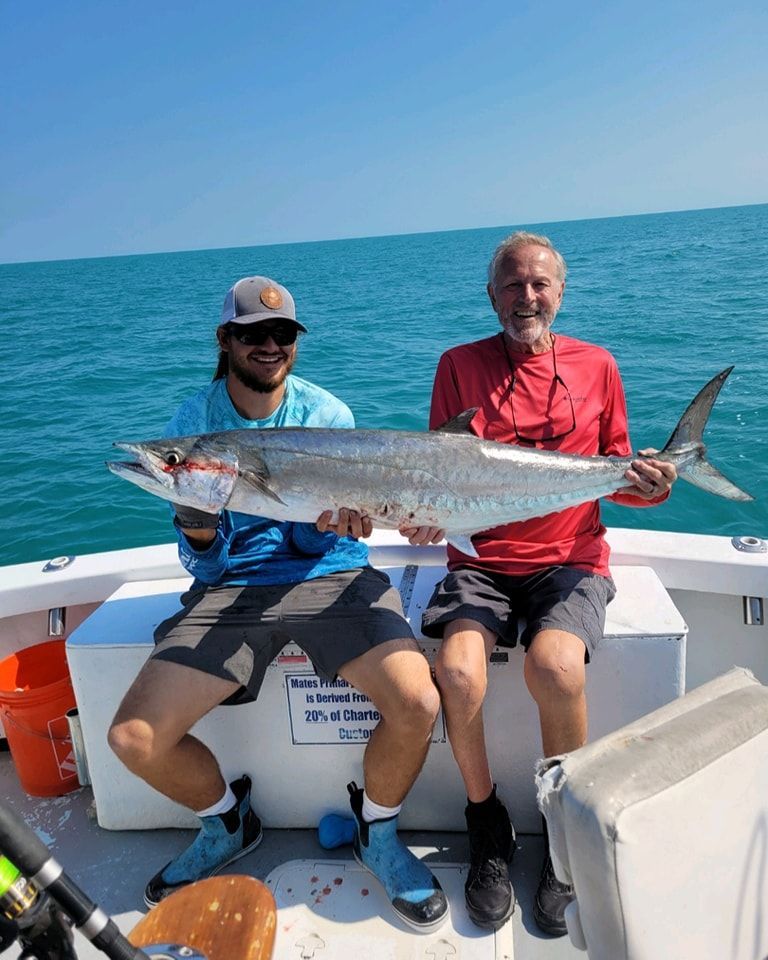 Two people sit on a boat deck holding a large, silvery fish caught in the open ocean under a clear blue sky.
