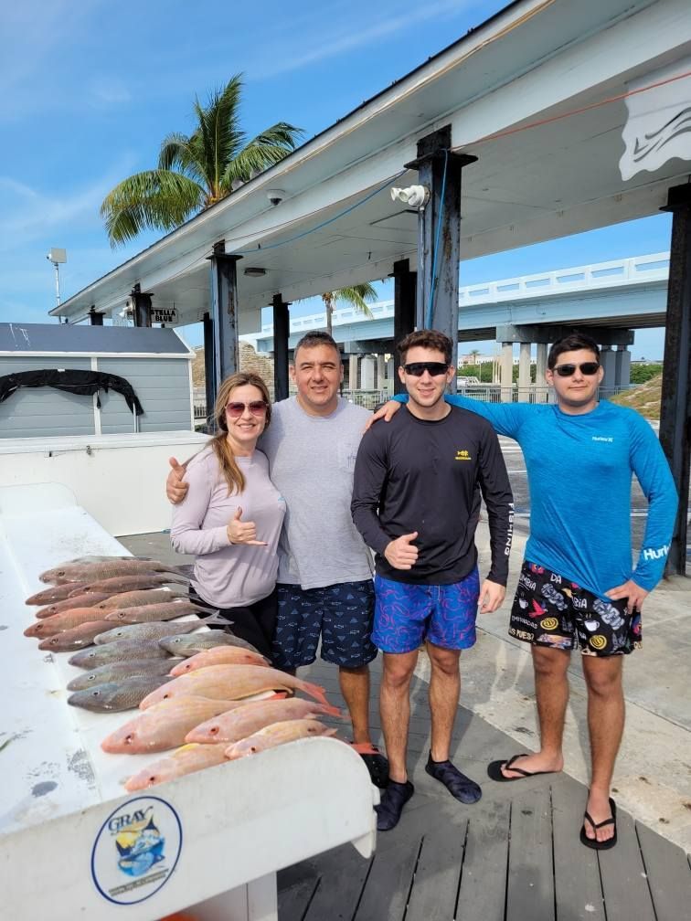 Four people stand smiling on a pier next to a table displaying several caught fish under a sunny sky.