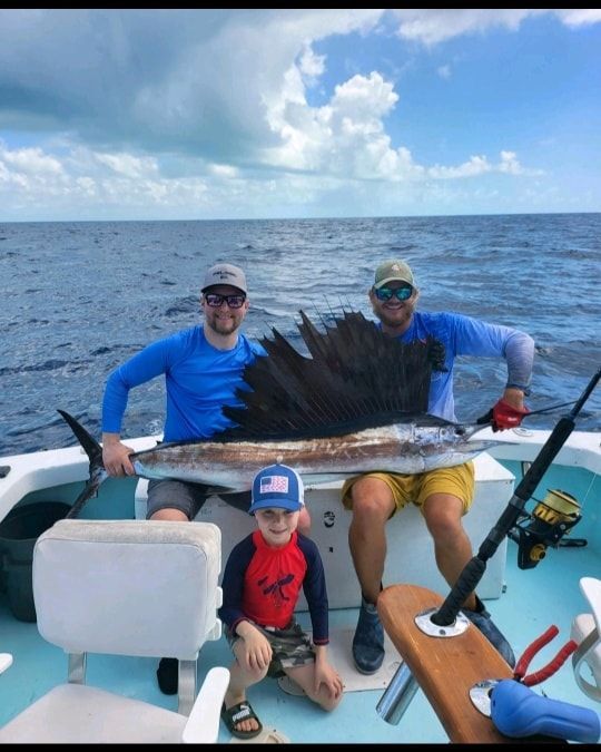 Two people and a child holding a large sailfish on the deck of a boat under a sunny sky.