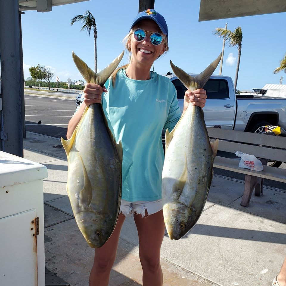 A smiling person holding two large fish by the tails while standing outdoors near a parked pickup truck.