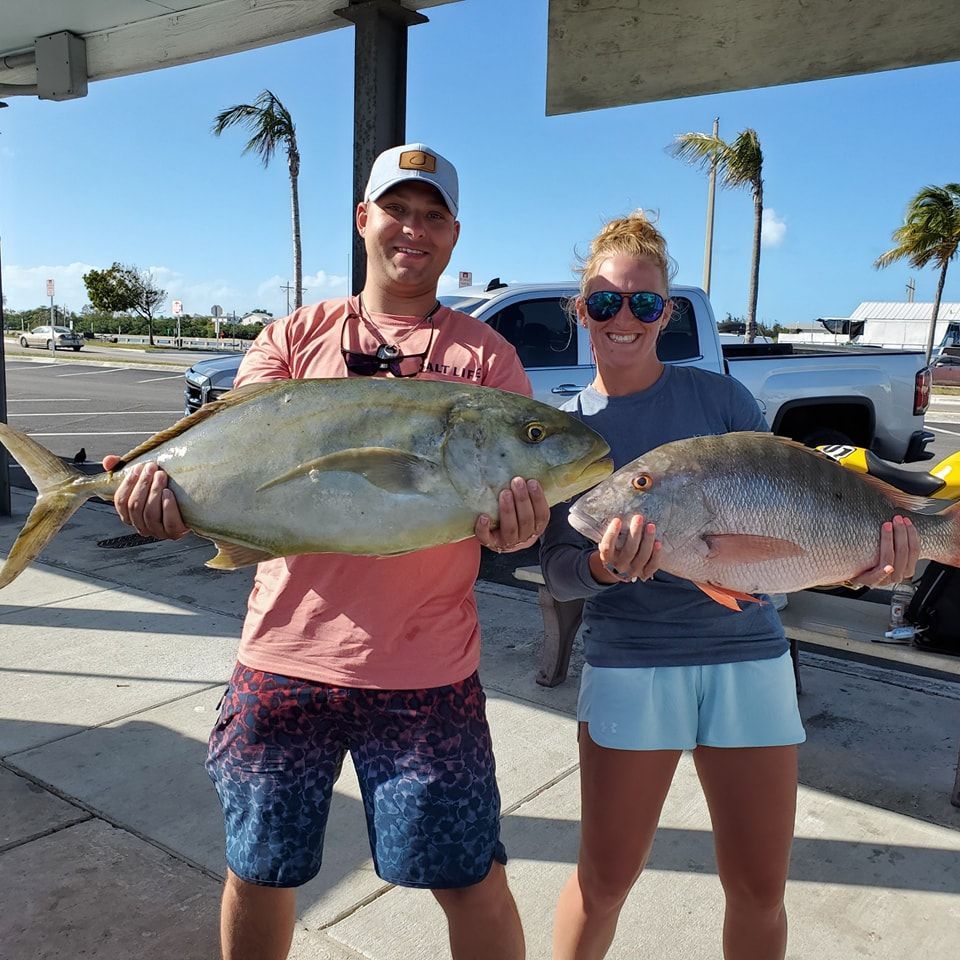 A man and woman standing outside hold two large fish they caught, with a white truck and palm trees in the background.
