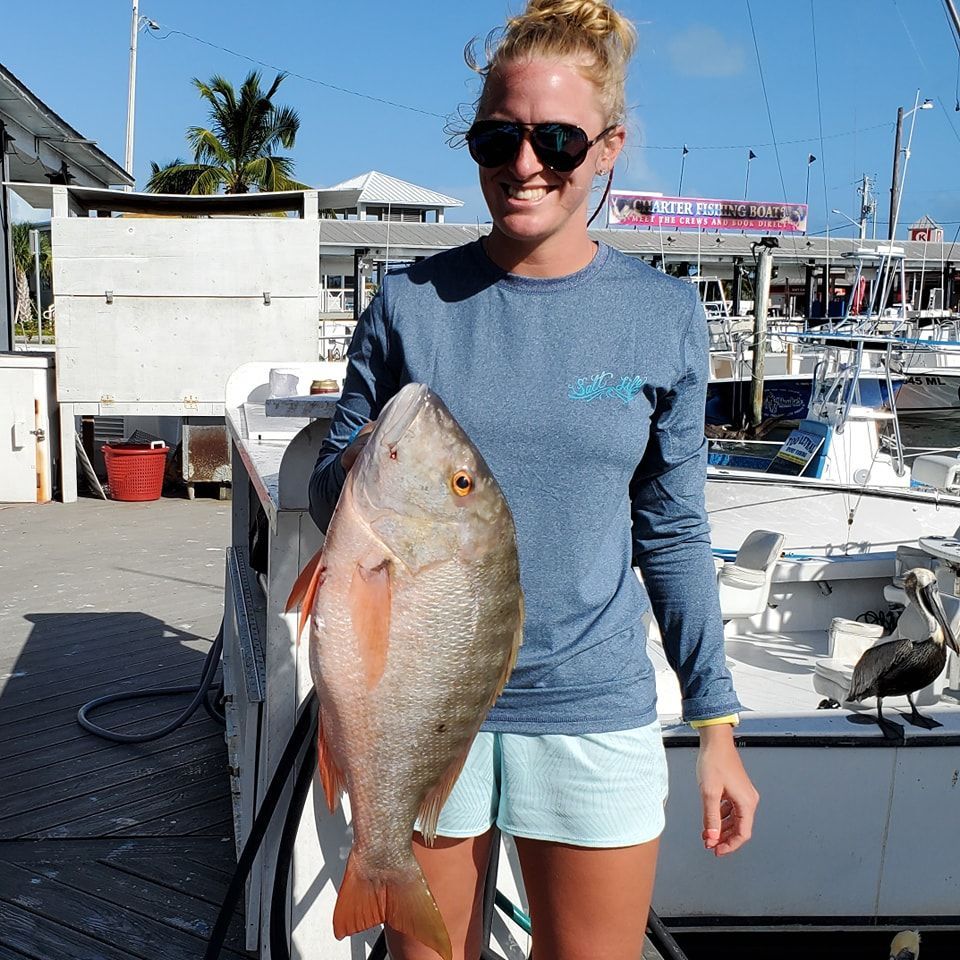 A person wearing sunglasses and a long-sleeved shirt holds a large, reddish-gray fish at a marina with boats and a pelican.