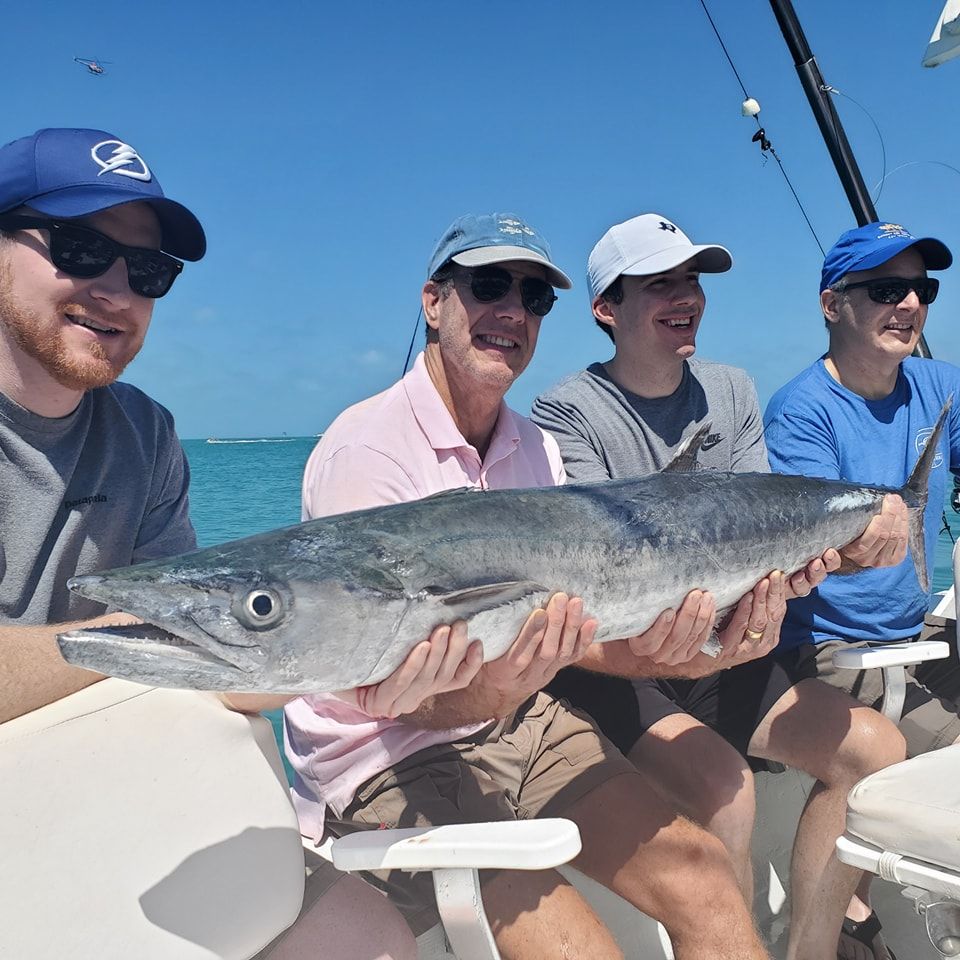 Four people on a boat smile while holding a large, silver fish horizontally between them against a clear blue sky.
