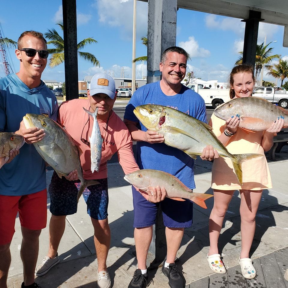Four people standing on a sunny pier, smiling while holding various types of freshly caught fish.