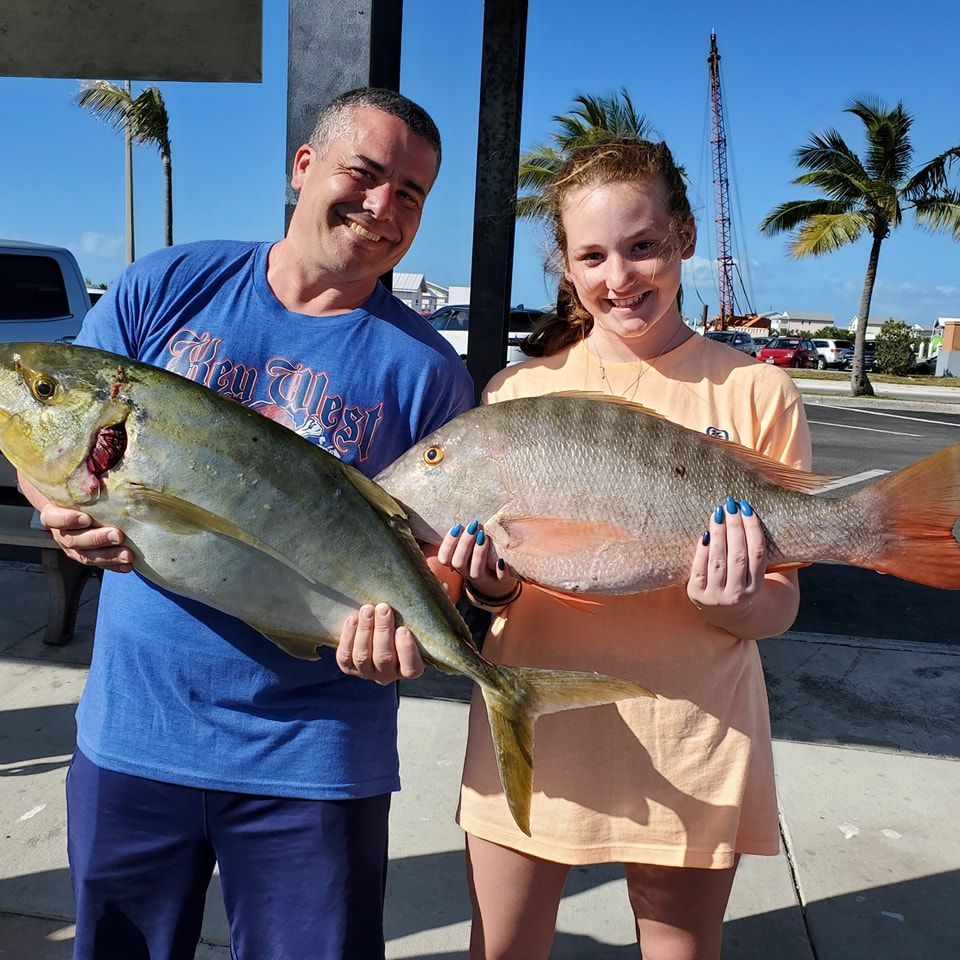 A man and a youth smile while holding two large, freshly caught fish in an outdoor parking area with palm trees.
