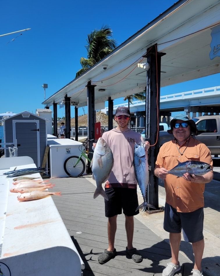 Two people hold large fish on a pier next to a cleaning station with several smaller fish lined up on a table.