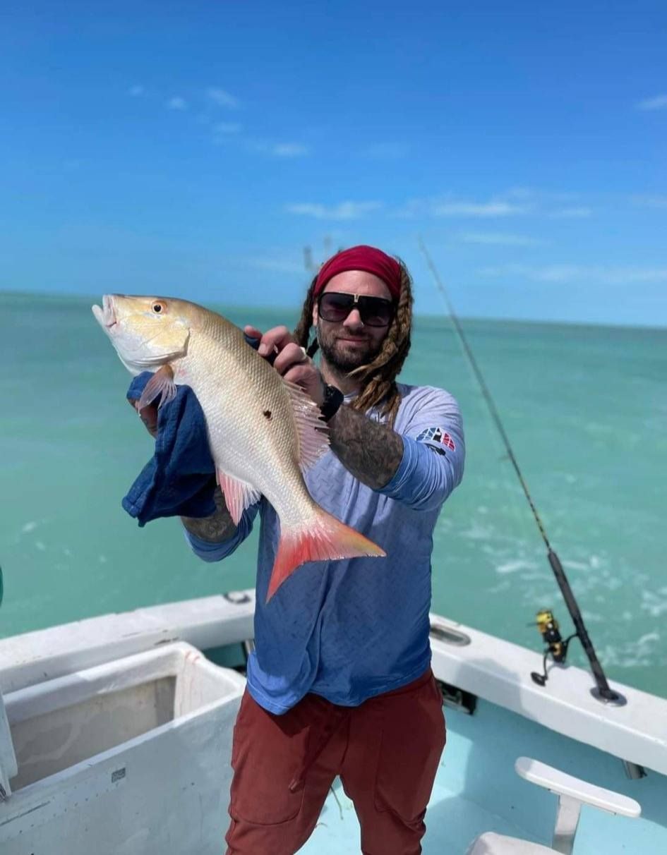 A person with dreadlocks and sunglasses holds a large red fish on a boat against a bright blue sky and ocean background.