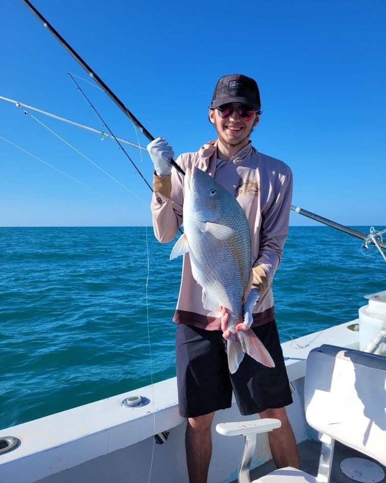 A person standing on a boat deck, smiling and holding up a large fish caught on a fishing line against a blue sea background.
