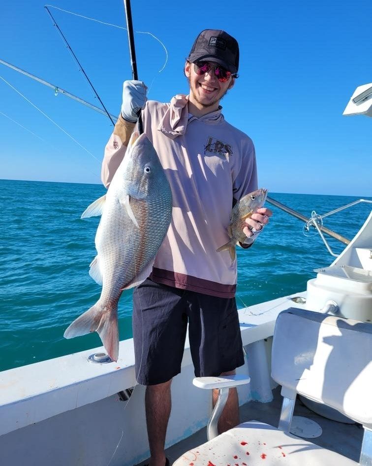 A person on a boat holds a large fish in one hand and a smaller fish in the other, smiling against a clear blue sky.
