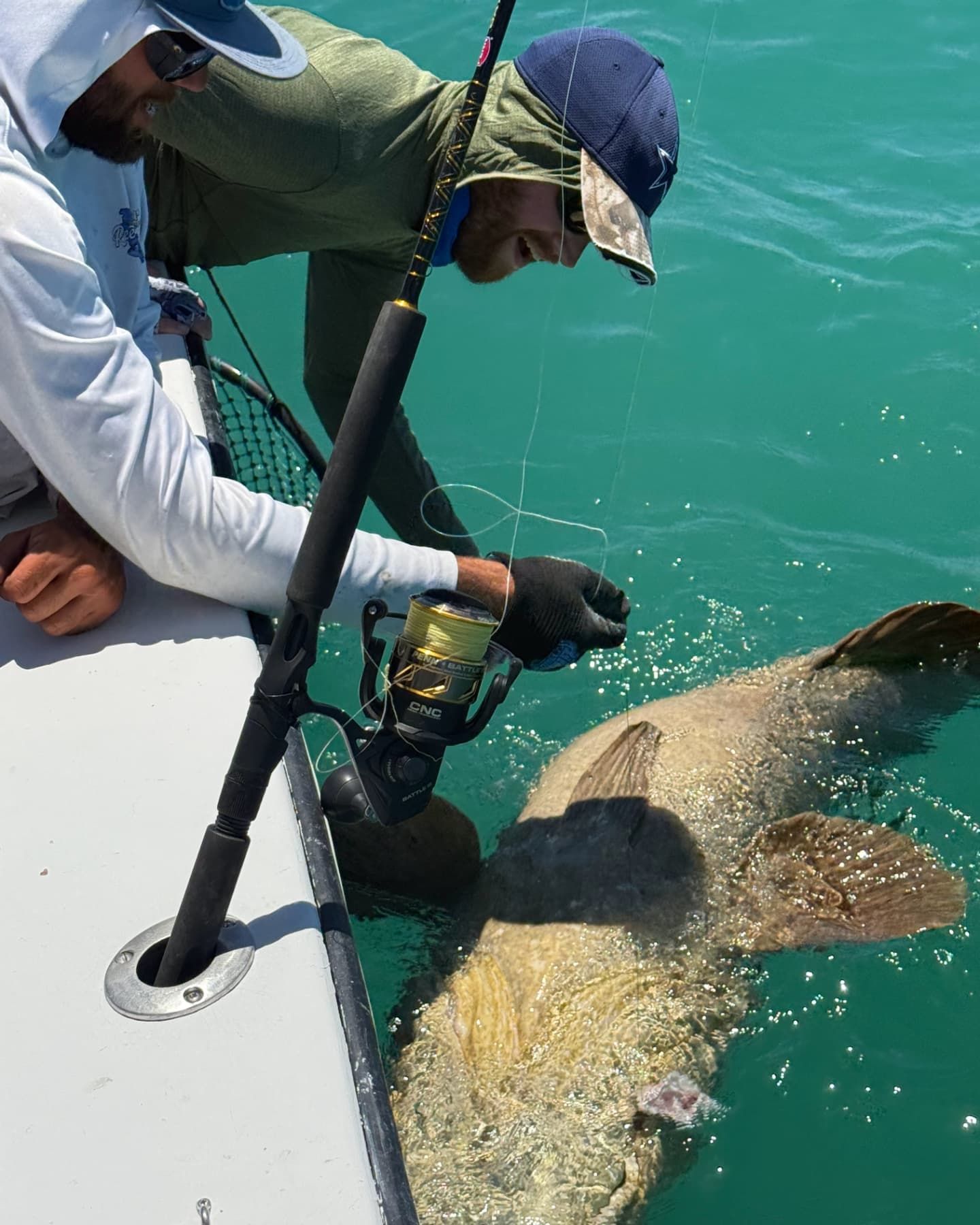 Two people on a boat in turquoise water hook a large, mottled fish near the surface.