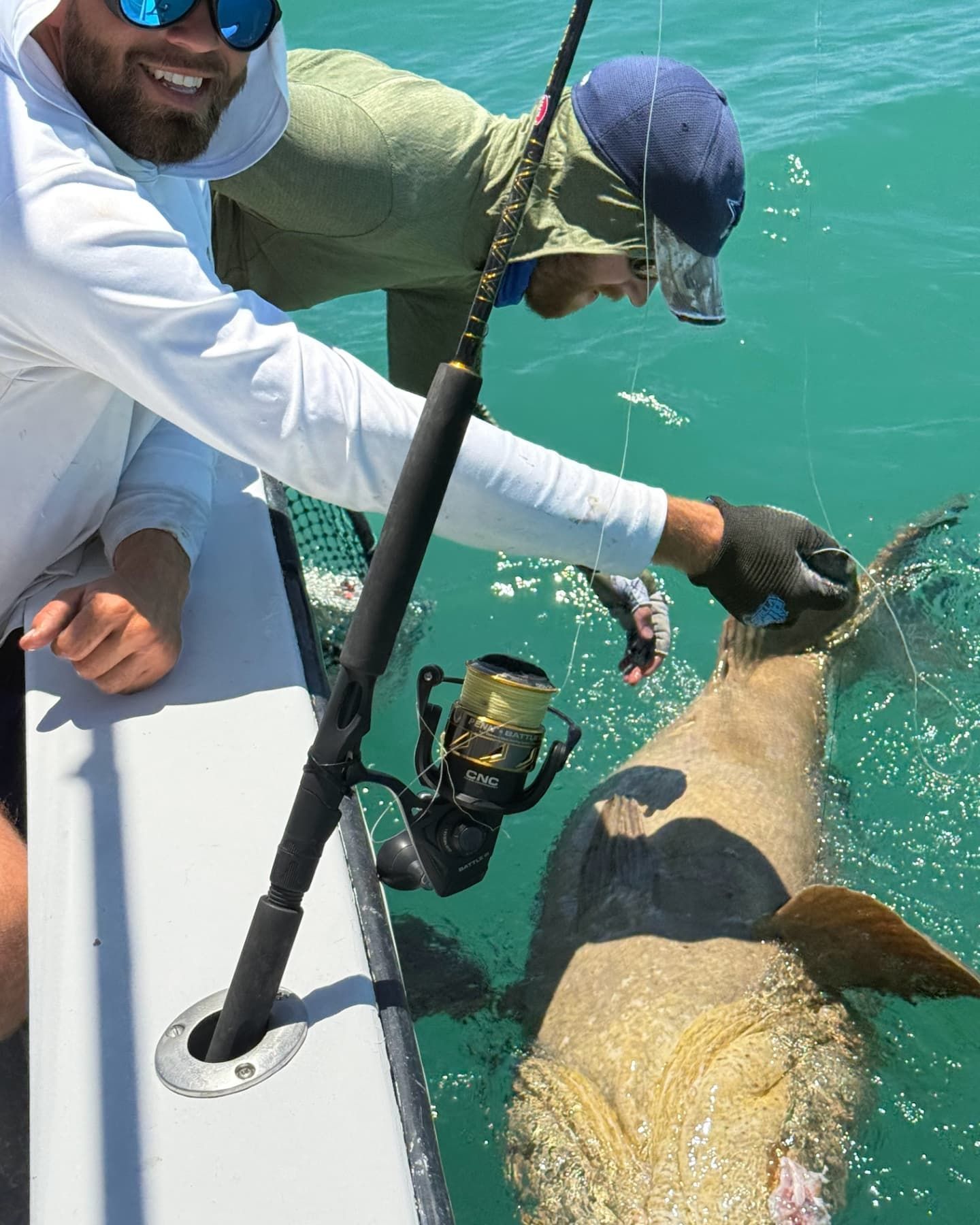 Two people on a boat in turquoise water reaching toward a large, light-colored fish near the surface.