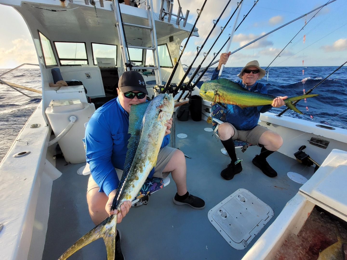Two people on a boat deck, smiling while each holding a large, bright yellow and green mahi-mahi fish after fishing.