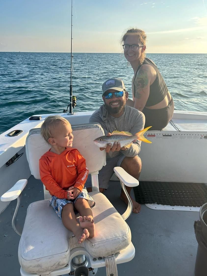 A family sits on a boat on the ocean, with a man holding a fish toward a young child sitting in a captain's chair.