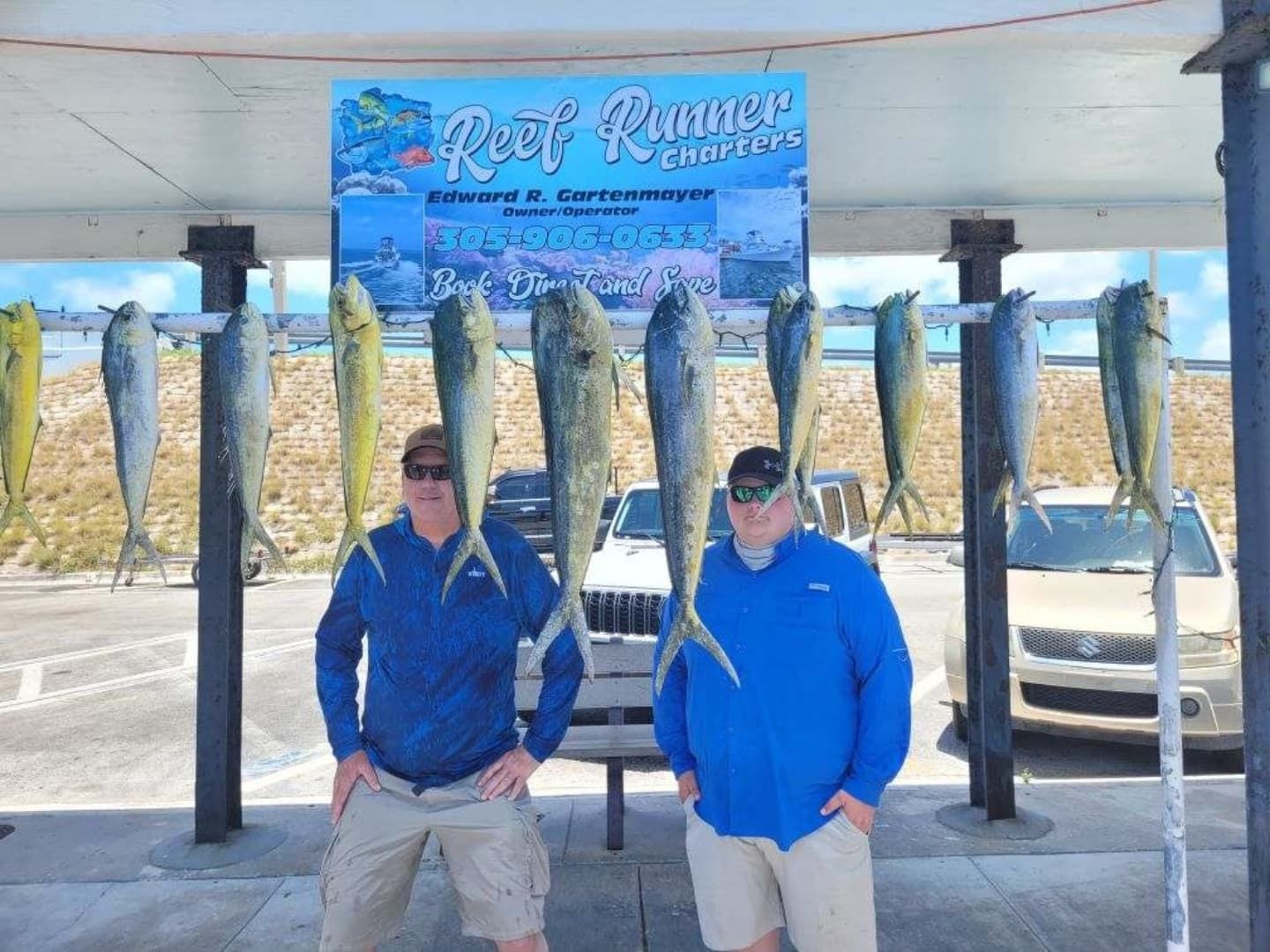 Two people in blue shirts stand outdoors posing with nine mahi-mahi fish hanging from a rack under a Reef Runner sign.