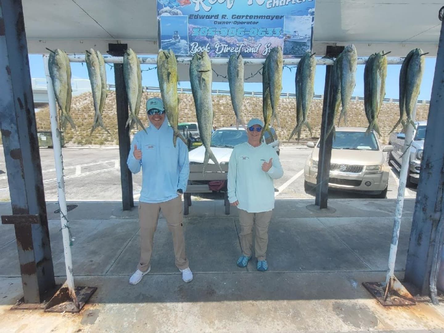 Two people stand smiling and giving thumbs-up under a rack displaying eight freshly caught mahi-mahi at a marina.