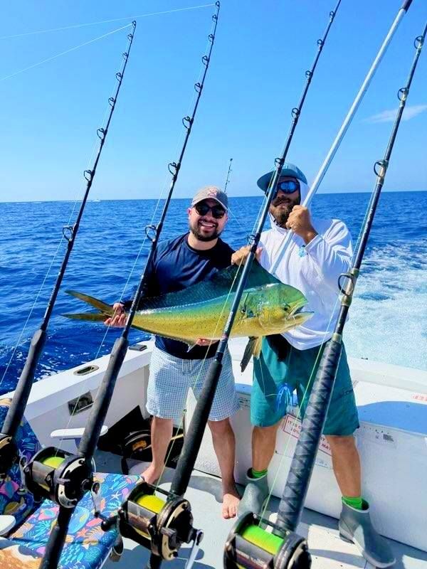 Two people on a boat hold a large, vibrant yellow and green mahi-mahi fish, surrounded by several tall fishing rods.