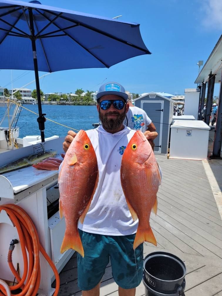 A person holds two freshly caught, bright orange snapper fish while standing on a dock under a blue umbrella.