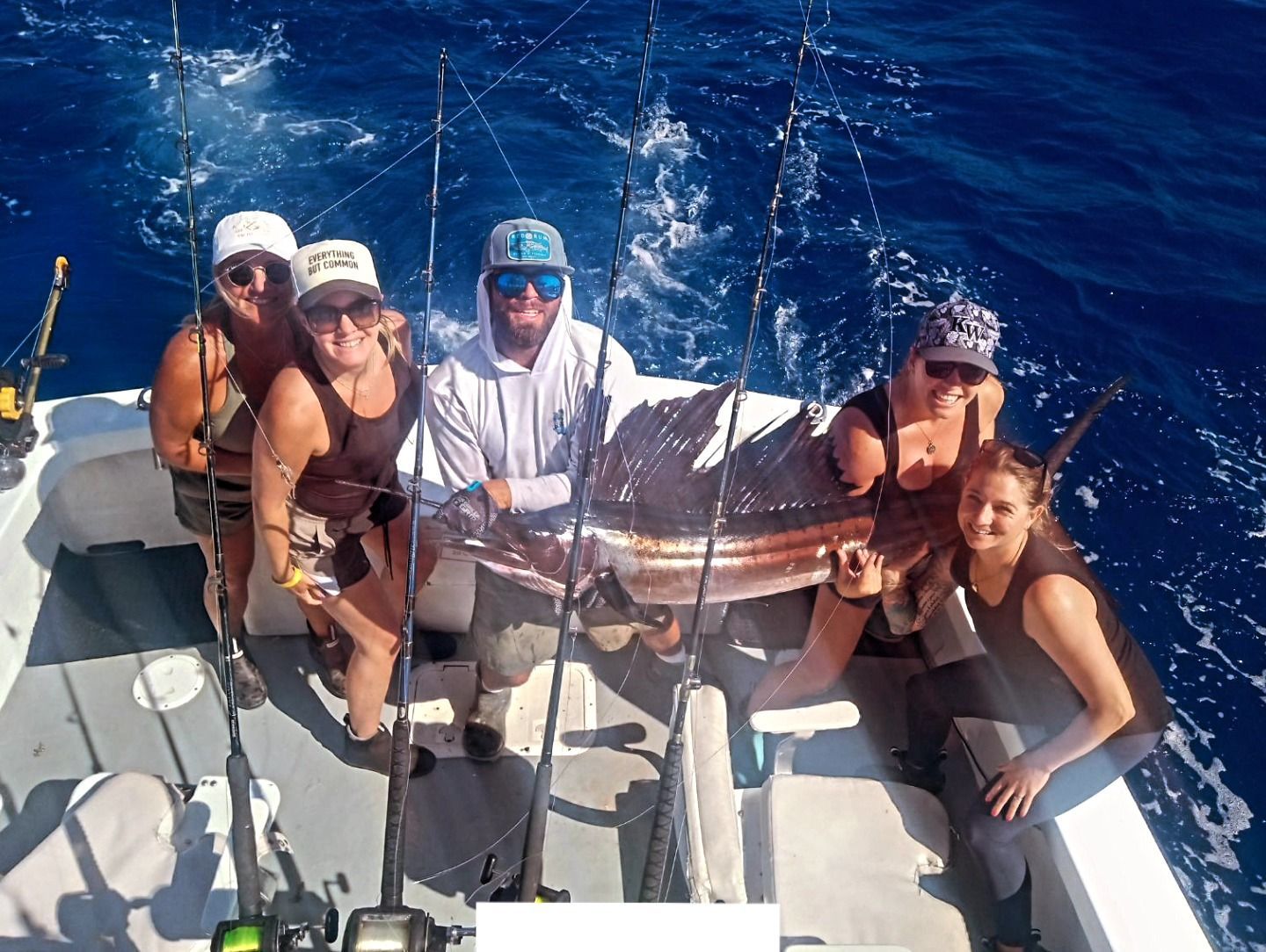 A group of five people on a boat pose with a large sailfish caught while fishing in the open ocean.