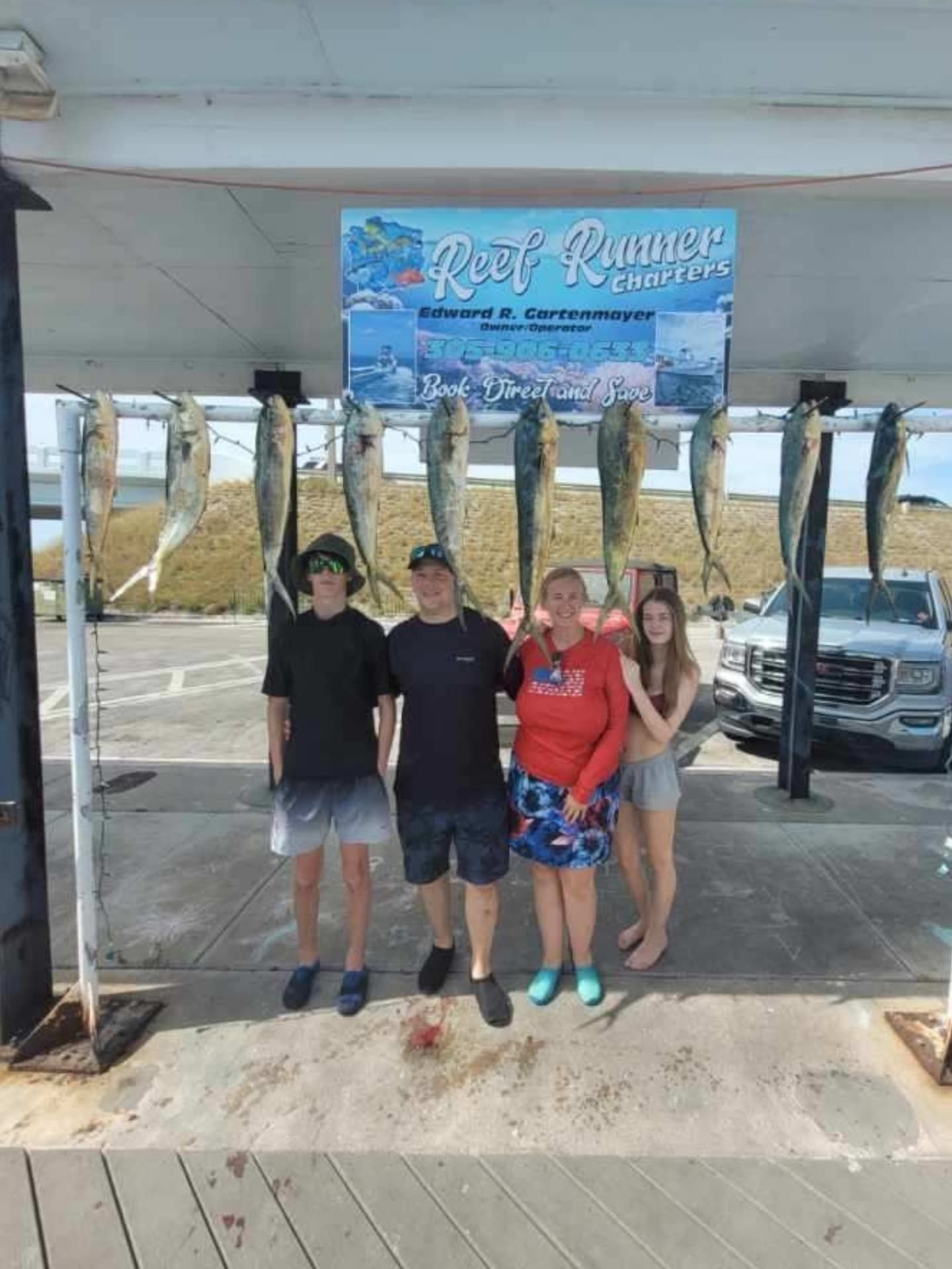 Four people stand smiling in front of a rack holding seven large, hanging fish at a Reef Runner charter boat facility.