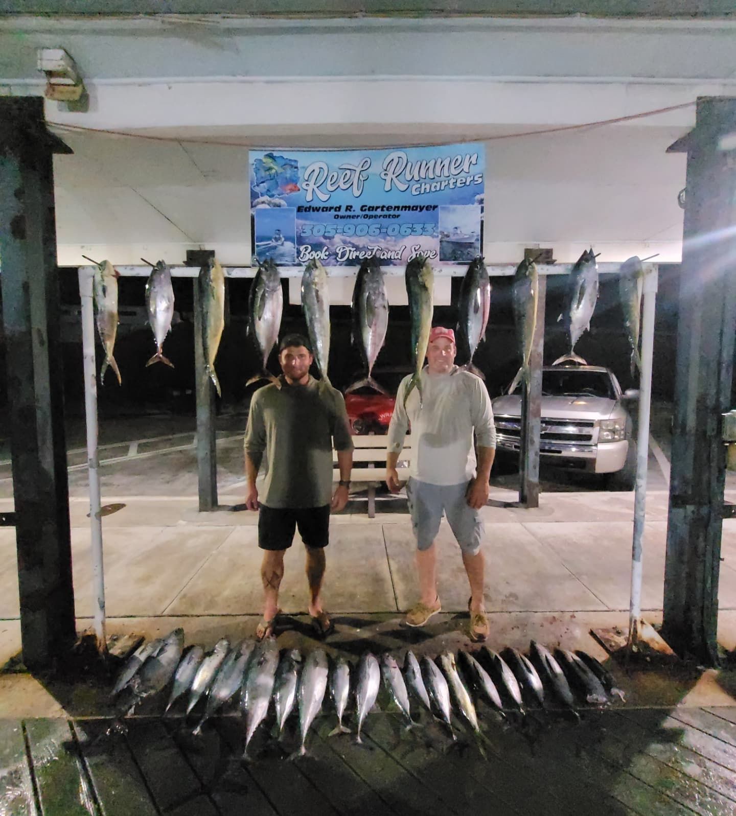 Two people stand in front of a sign and a rack holding many hanging fish, with more fish arranged on the floor below.