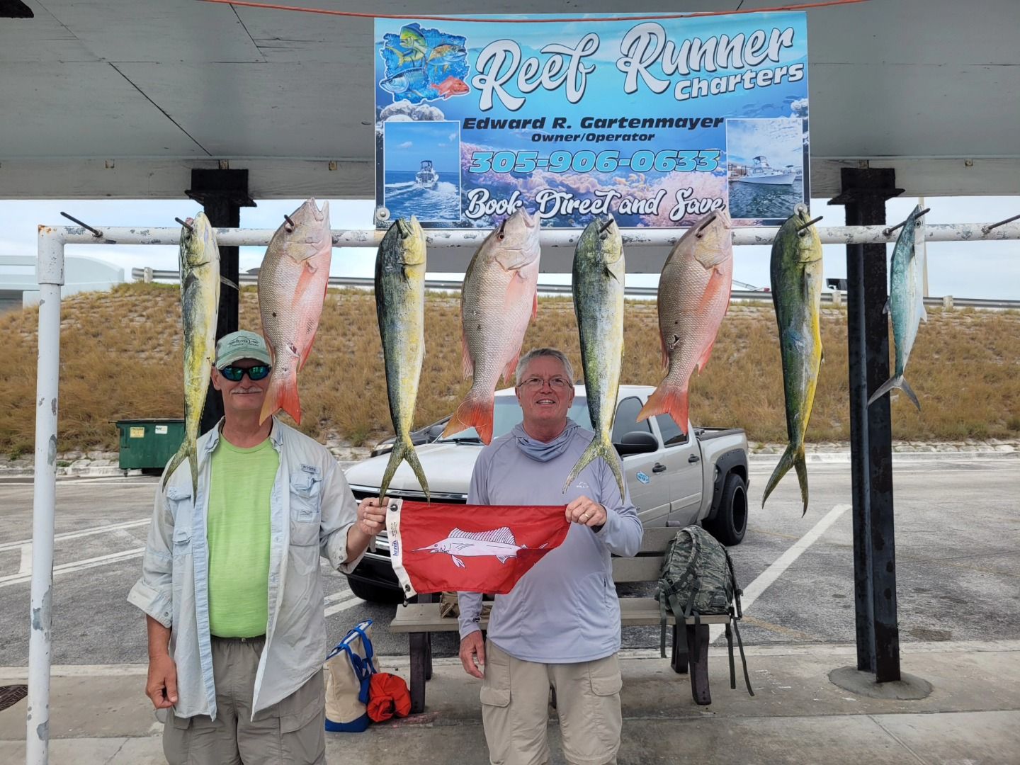 Two people stand with caught fish hanging on a rack in front of a Reef Runner Charter boat sign at a marina.