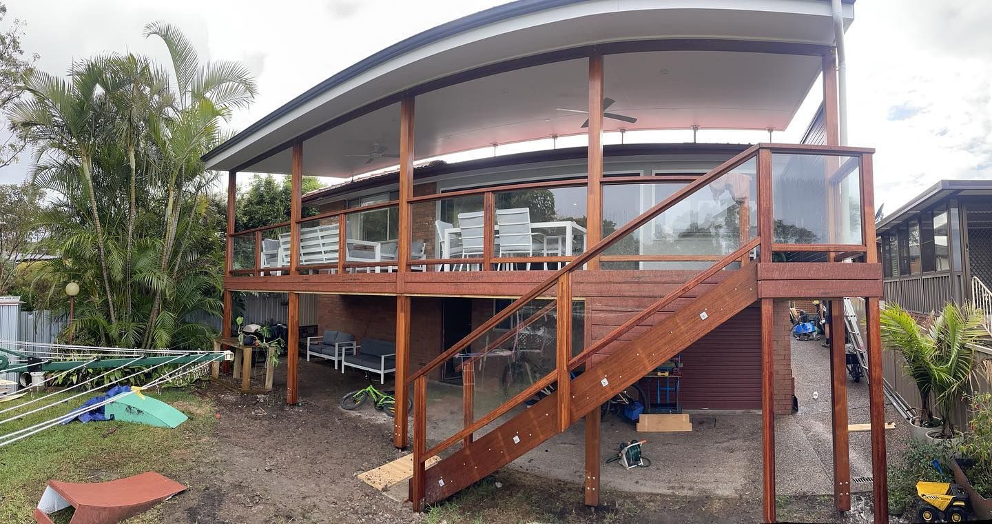 Wooden house with a two-story deck, glass railings, and a curved roof overlooking a yard with greenery.