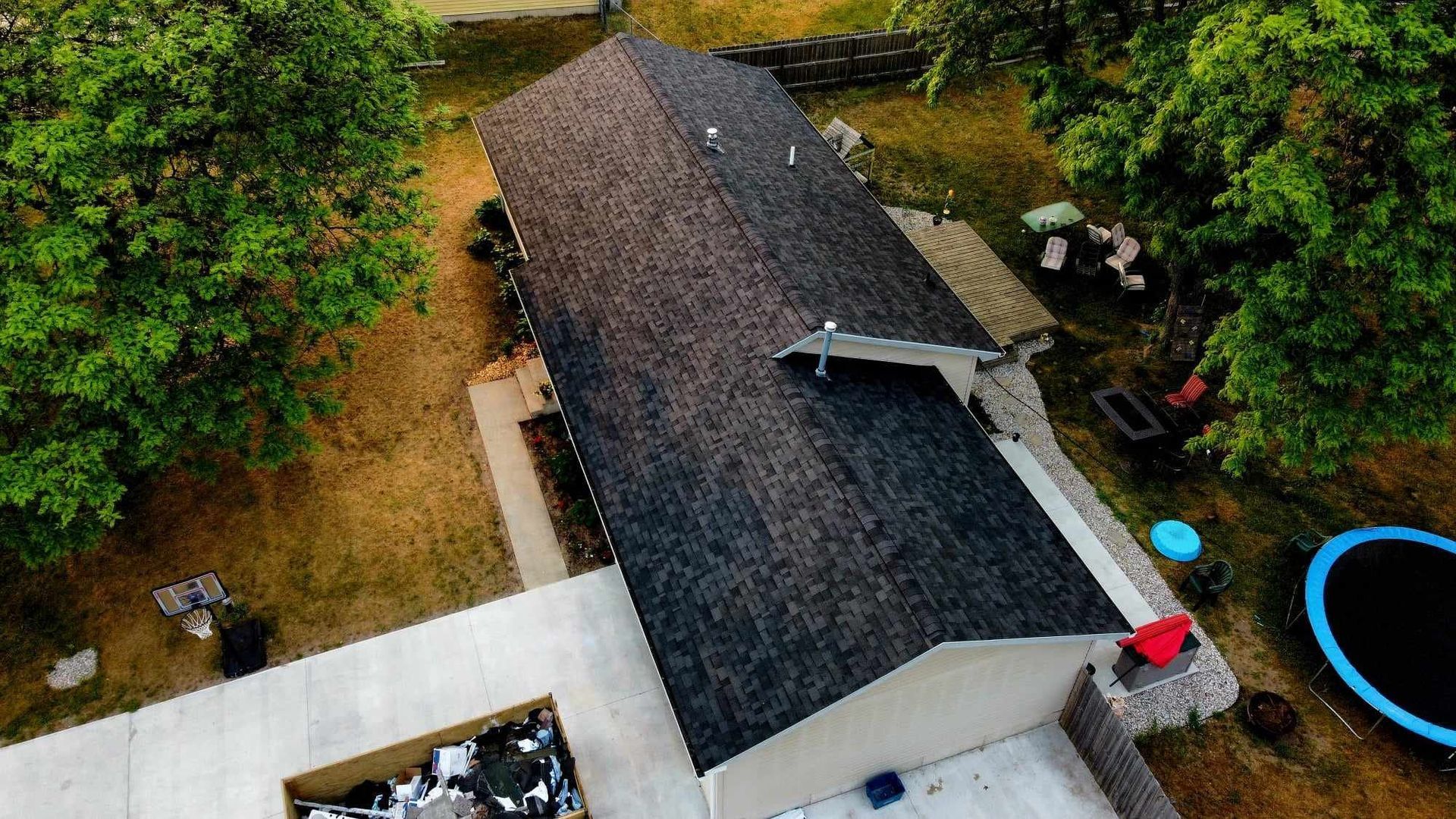 An aerial view shows a new dark gray shingled roof on a home, with a construction dumpster and a backyard trampoline nearby.